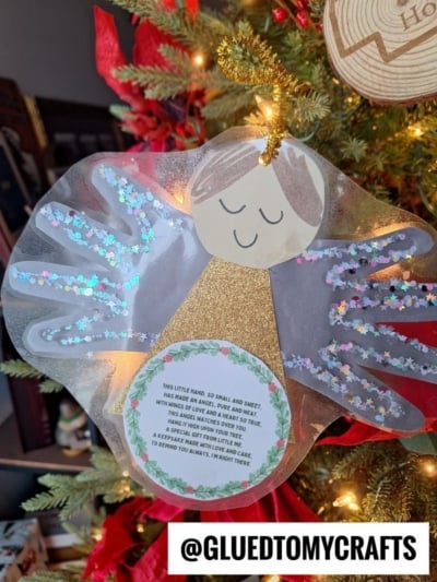 A handmade handprint angel ornament with glittery wings and a smiling face hangs on the Christmas tree. A heartfelt poem is attached, and a wooden ornament is visible in the background. @gluedtomycrafts is at the bottom.