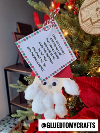 A handmade handprint Santa ornament with googly eyes hangs on a Christmas tree. A festive tag reads, Look very close! That isn’t Santa’s beard—hang this ornament on your tree and share the joy with the whole family!.