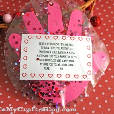 A handmade craft featuring a pink paper handprint with red glitter, a my little handprint poem card attached, and heart decorations, displayed on a red polka-dot background
