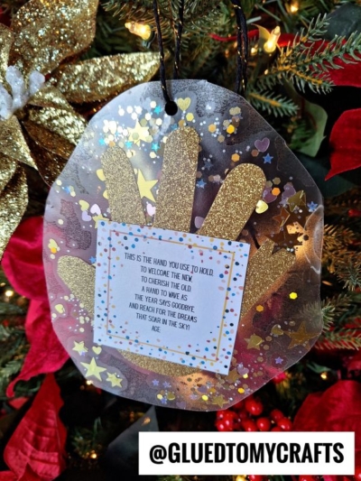 A handmade ornament hangs on a Christmas tree, featuring a gold new year's eve handprint, colorful confetti, and a poem about cherishing the old year and welcoming the new. Decorative gold leaves and red poinsettias are visible in the background.