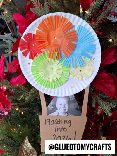 A paper plate hot air balloon craft—perfect for New Year’s Eve Crafts For Kids—decorated with colorful paper flowers and gold sequins, features a smiling child's photo and a “Floating into 2024!” sign, displayed on a Christmas tree.