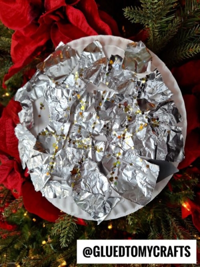 A round paper plate covered with crumpled aluminum foil and sprinkled with gold glitter is displayed against red poinsettias and green pine branches—a festive idea for New Year’s Eve Crafts For Kids. A tag reads @GLUEDTOMYCRAFTS.