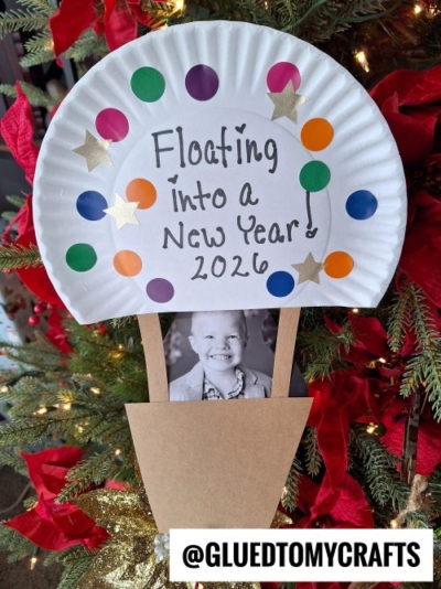A paper plate craft resembling a hot air balloon is decorated with colorful dots and stars, reading Floating into a New Year 2026! Perfect for New Year’s Eve Crafts For Kids, it features a black-and-white child’s photo as the basket. @GLUEDTOMYCRAFTS is written at the bottom.
