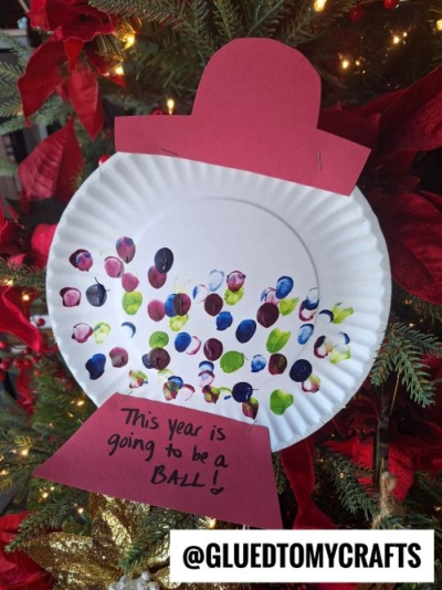 A paper plate snow globe craft with colorful thumbprint dots and a red paper base, hanging on a Christmas tree. A handwritten note reads, This year is going to be a ball!—a fun idea for New Year's Eve crafts by @GLUEDTOMYCRAFTS.
