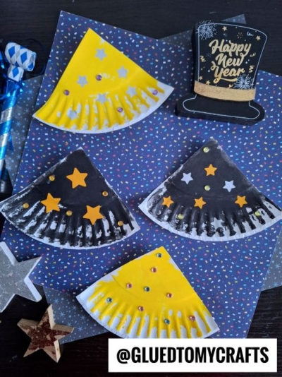 Paper plate crafts painted yellow and black, decorated with stars and sequins, are arranged on a festive dotted background. Perfect for New Year’s Eve Crafts For Kids! A Happy New Year sign and party items add extra cheer. Text at bottom reads @GLUEDTOMYCRAFTS.