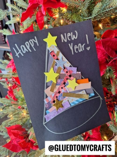 A handmade New Year’s card, perfect for New Year’s Eve Crafts For Kids, features a collage Christmas tree from colorful paper scraps and star stickers on black paper. “Happy New Year!” is written in silver marker, with a festive tree in the background.