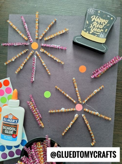 A New Year’s fireworks craft with colorful pipe cleaners arranged as fireworks on black paper, orange dot stickers, glue, and a Happy New Year hat cutout—perfect inspiration for New Year’s Eve Crafts For Kids. @GLUEDTOMYCRAFTS is written at the bottom.