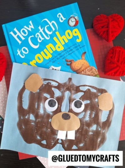 A paint splat groundhog craft—featuring brown paint, paper shapes, and googly eyes—is displayed in front of the book How to Catch a Groundhog. Red heart decorations add a festive touch in the background.