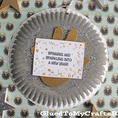 A silver paper plate disco ball decorated with glitter, a gold cutout, and a card that reads “Spinning and sparkling into a new year!” is surrounded by clock and star motifs.