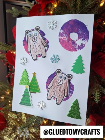 A holiday craft page with colored bears, green Christmas tree cutouts, glittery snowflakes, and a pool noodle stamped snow tube, plus purple circular stamps, all displayed in front of a decorated Christmas tree. @GLUEDTOMYCRAFTS is written at the bottom.