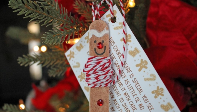 A popsicle stick decorated as a gingerbread man ornament with googly eyes, red nose, buttons, and a scarf, holding a holiday card near a Christmas tree.
