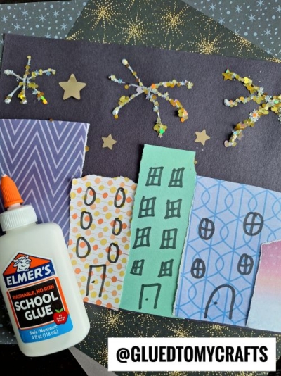 A colorful cityscape craft made with patterned paper buildings, a black background with star and firework decorations—perfect for New Year’s Eve Crafts For Kids—plus gold star stickers, glitter accents, and a bottle of Elmer’s glue. @GLUEDTOMYCRAFTS is at the bottom.