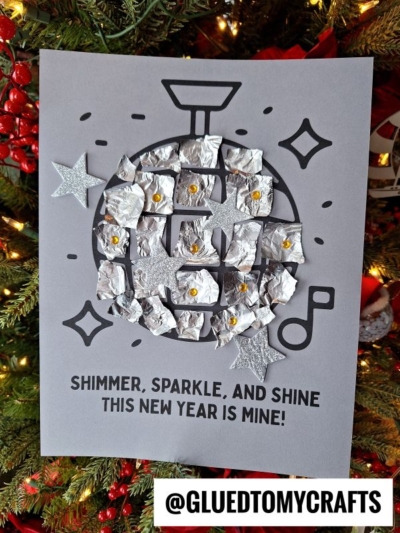A gray paper with a black torn paper disco ball outline, decorated with crumpled foil and glittery stars. Below, text reads, Shimmer, sparkle, and shine. This New Year is mine! The background features a Christmas tree.
