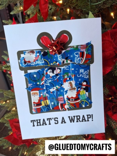 A Christmas card shaped like a torn paper present, decorated with blue Santa-themed wrapping paper, a shiny red bow on top, and the words “THAT’S A WRAP!” at the bottom, displayed in front of a Christmas tree.
