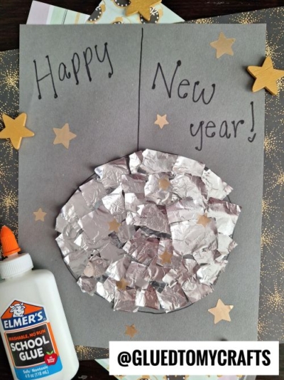 A handmade New Years card with Happy New Year! on gray paper, a shiny silver collage circle, gold star cutouts, a bottle of Elmers glue—perfect for New Year’s Eve Crafts For Kids. The text @GLUETOMYCRAFTS is at the bottom.