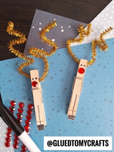 Two clothespins decorated as reindeer with gold pipe cleaner antlers, red gem noses, and drawn eyes. Perfect for CHRISTMAS CRAFTS FOR KIDS! Craft supplies like red gems, a marker, and blue star paper are nearby. @GLUEDTOMYCRAFTS is at the bottom.