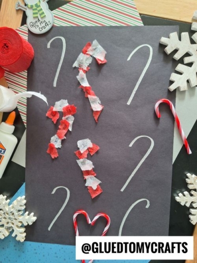 A black paper with white candy cane outlines, some filled with red and white tissue, is surrounded by craft supplies, candy canes, snowflake cutouts, and a @GLUEDTOMYCRAFTS watermark—perfect inspiration for CHRISTMAS CRAFTS FOR KIDS.
