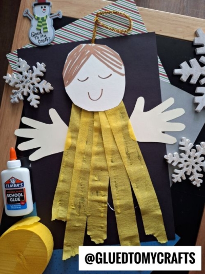 A handcraft angel made from paper with a round head, drawn face, yellow crepe streamer body, and paper hands—perfect for CHRISTMAS CRAFTS FOR KIDS—surrounded by craft supplies, snowflake decorations, and an @GLUEDTOMYCRAFTS label.