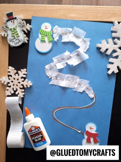 A blue paper with a wavy path made from torn white tissue, surrounded by wooden snowflakes, snowman stickers, a glue bottle, and a roll of white streamer—perfect inspiration for CHRISTMAS CRAFTS FOR KIDS. Text reads @GLUEDTOMYCRAFTS at the bottom.