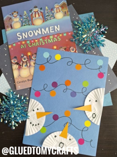 A children’s book titled Snowmen at Christmas is next to a snowman craft made with blue paper, cupcake liners, colored paper circles, and marker details—perfect inspiration for Activities For Kids To Do At Home. Sparkly blue snowflakes decorate the background.
