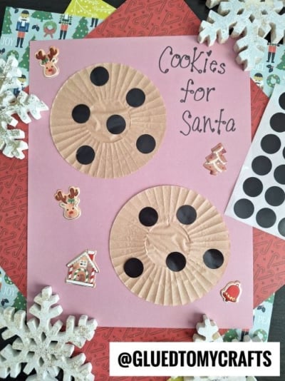 A pink paper with two cupcake liners decorated as chocolate chip cookies, labeled Cookies for Santa, surrounded by festive stickers and white snowflake ornaments—perfect CHRISTMAS CRAFTS FOR KIDS with black dot stickers and craft supplies nearby.