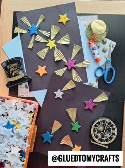 Colorful New Year’s Eve craft supplies for kids on a table, featuring black paper with glitter stars, gold cupcake liner fireworks, glue stick, scissors, a clock cutout, and foam stars. Perfect inspiration after your CHRISTMAS CRAFTS FOR KIDS fun! @GLUEDTOMYCRAFTS.