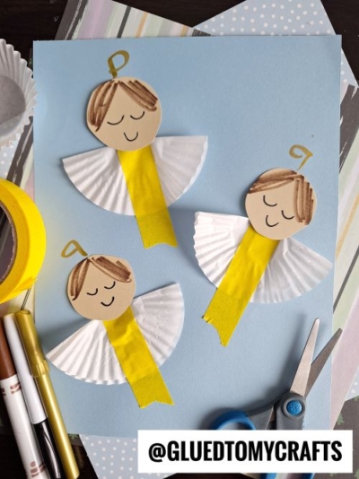 Three paper angels made from cupcake liners, yellow ribbon, and paper circles with drawn faces are arranged on a light blue background—perfect CHRISTMAS CRAFTS FOR KIDS. Craft supplies like markers, scissors, and tape are nearby.