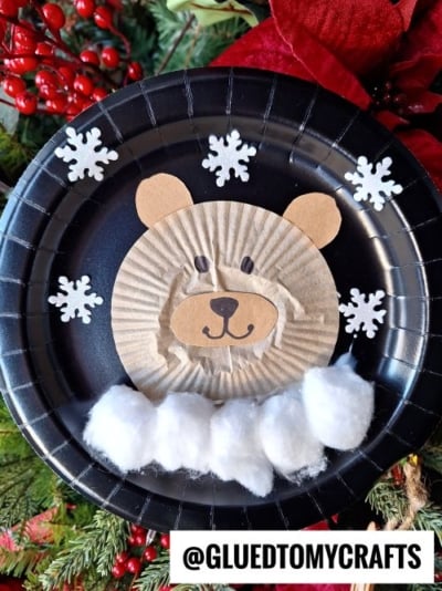A bear face made from a tan cupcake liner and paper sits on a black plate, surrounded by white snowflakes and cotton balls, with red berries and greenery behind—perfect inspiration for CHRISTMAS CRAFTS FOR KIDS. @GLUEDTOMYCRAFTS is at the bottom.