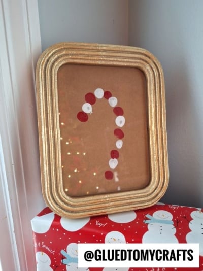 A gold-framed picture with a candy cane shape made of red and white paint dots, displayed on a snowman-patterned wrapped box. Perfect for CHRISTMAS CRAFTS FOR KIDS. The handle @GLUEDTOMYCRAFTS is shown at the bottom.