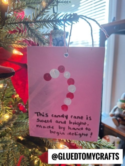 A handmade card ornament with red and white buttons forming a candy cane hangs on a Christmas tree. Perfect for CHRISTMAS CRAFTS FOR KIDS, it reads, This candy cane is sweet and bright, made by hand to begin delight! @GLUEDTOMYCRAFTS is printed at the bottom.