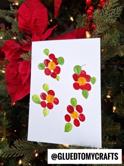 A white card with colorful fingerprint art of red flowers and green leaves, perfect for CHRISTMAS CRAFTS FOR KIDS, is displayed on a Christmas tree with red ribbon and ornaments. Text at the bottom reads @GLUEDTOMYCRAFTS.