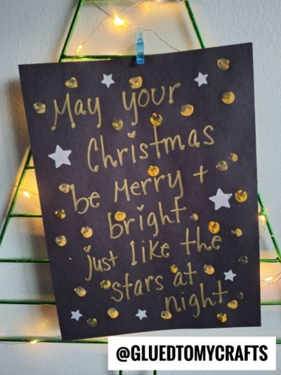 A black paper sign with gold writing and white stars reads: “May your Christmas be Merry + Bright, just like the stars at night.” Perfect for CHRISTMAS CRAFTS FOR KIDS, the sign is clipped to a green wire frame with lights. @GLUEDTOMYCRAFTS appears at the bottom.
