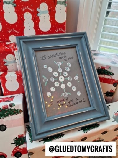 A framed craft with a snowflake made of white fingerprints and the words “If snowflakes were kisses would you want a blizzard?” sits in front of holiday-themed gift boxes—perfect inspiration for Christmas crafts for kids.