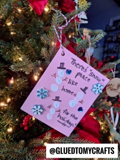 A handmade pink card hangs on a Christmas tree, perfect for CHRISTMAS CRAFTS FOR KIDS. It features snowmen made from white buttons, blue snowflake decorations, and reads "There's snow place like home for the holidays!.