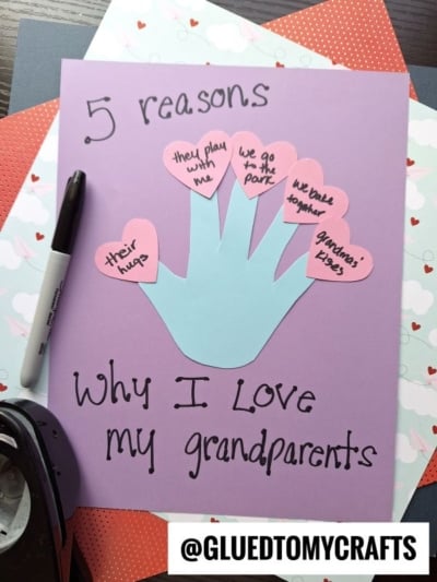 A purple paper features a blue valentine's day handprint cutout, each finger topped with a pink heart listing reasons for loving grandparents, like “their hugs” and “we go to the park.” Marker, hole punch, and papers sit nearby.
