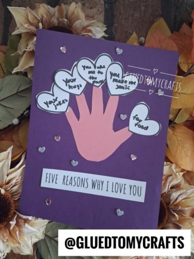 A purple card with a pink valentine’s day handprint cutout, each finger labeled with a reason for love. Surrounded by heart shapes and sparkly stickers, the card says FIVE REASONS WHY I LOVE YOU on a floral background.