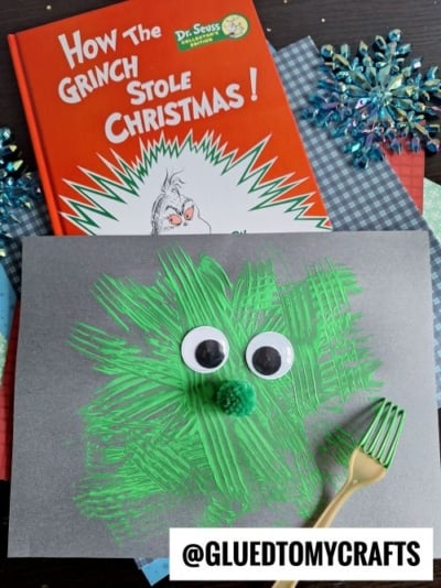 A festive CHRISTMAS CRAFTS FOR KIDS project inspired by How the Grinch Stole Christmas! uses green paint textured with a fork, two jumbo googly eyes, and a green pompom nose on gray paper, alongside the book and craft supplies in the background.