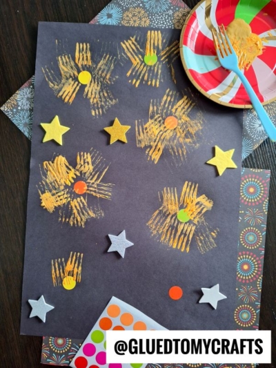 A black sheet with yellow painted starbursts, foam star stickers, and colored dot stickers—perfect for New Year’s Eve Crafts For Kids. A plate with a partially eaten cookie and a plastic fork sits nearby. The handle @GLUEDTOMYCRAFTS is at the bottom.