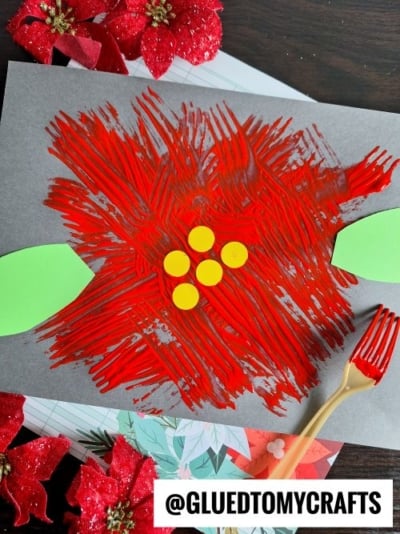 A child’s craft of a bright red poinsettia flower painted with fork strokes on dark paper, featuring green paper leaves and yellow paper circles as the flower’s center—perfect for CHRISTMAS CRAFTS FOR KIDS, with a red plastic fork and red flowers nearby.