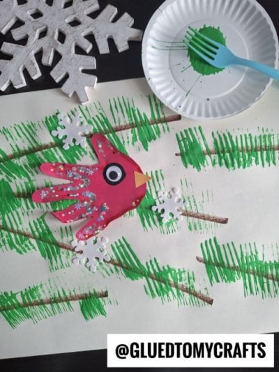 A fun CHRISTMAS CRAFTS FOR KIDS project features a red bird made from a cut-out handprint, decorated with white dots and a googly eye, perched on green-painted pine branches. White paper snowflakes and a plate with green paint complete the scene.