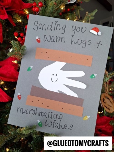 A handmade holiday card, perfect for CHRISTMAS CRAFTS FOR KIDS, features a cutout white handprint, brown paper s’mores, and the message “Sending you warm hugs + marshmallow wishes,” finished with festive lights and a tiny Santa hat.