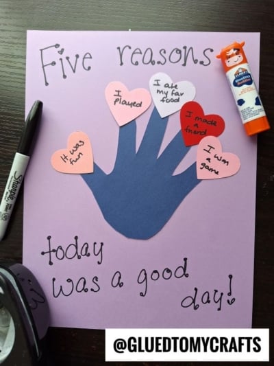 A purple paper with a blue valentine's day handprint cutout and five heart shapes listing reasons for a good day, surrounded by a marker, glue stick, and hole punch. The text reads Five reasons today was a good day! and @GLUEDTOMYCRAFTS.