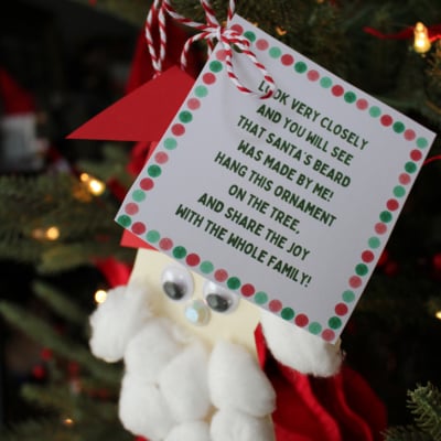 A handmade handprint Santa ornament with a cotton ball beard and googly eyes hangs on a Christmas tree. A card reads, “Look very closely and you will see that Santa’s beard was made by me!” Red ribbon and lights decorate the tree.