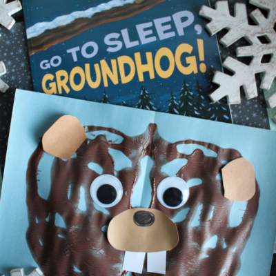 A fun children's craft project featuring a paint splat groundhog face with googly eyes, paper ears, nose, and teeth on blue paper, placed near the book Go to Sleep, Groundhog! and decorated with snowflakes.