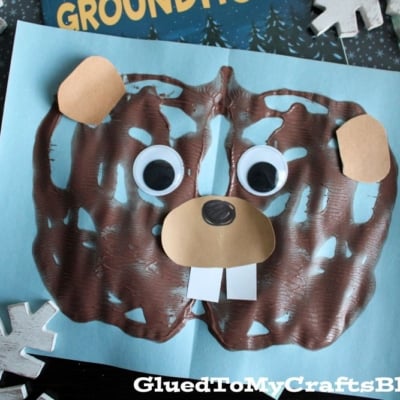 A fun children's craft project featuring a paint splat groundhog face with googly eyes, paper ears, nose, and teeth on blue paper, placed near the book Go to Sleep, Groundhog! and decorated with snowflakes.