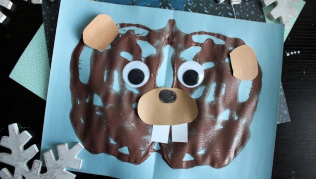 A fun children's craft project featuring a paint splat groundhog face with googly eyes, paper ears, nose, and teeth on blue paper, placed near the book Go to Sleep, Groundhog! and decorated with snowflakes.