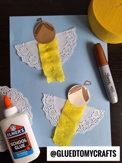 Two angel crafts made from paper doilies, yellow ribbon, and brown paper circles with hair details are displayed on blue paper—an adorable idea for CHRISTMAS CRAFTS FOR KIDS. Nearby are school glue, a black Sharpie, and yellow crepe paper.