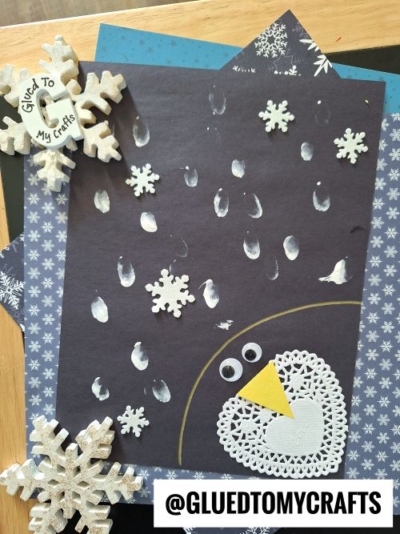 A winter-themed CHRISTMAS CRAFTS FOR KIDS project with a black sheet featuring white fingerprint snowflakes, a paper snowflake owl face with googly eyes and yellow beak, plus decorative snowflakes. @GLUEDTOMYCRAFTS is written at the bottom.
