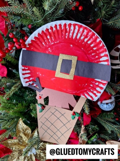 A Christmas tree decorated with a handmade paper plate Santa craft and a reindeer in a paper sleigh—perfect for CHRISTMAS CRAFTS FOR KIDS. The Santa face is made from a red plate with a black and gold belt. @GLUEDTOMYCRAFTS is written at the bottom.