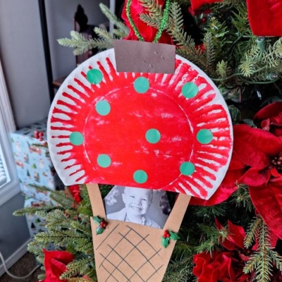 A handmade Christmas ornament, perfect for CHRISTMAS CRAFTS FOR KIDS, is shaped like an ice cream cone with a red and white top and green dots. It hangs on a decorated tree and features a small black-and-white photo of a child.